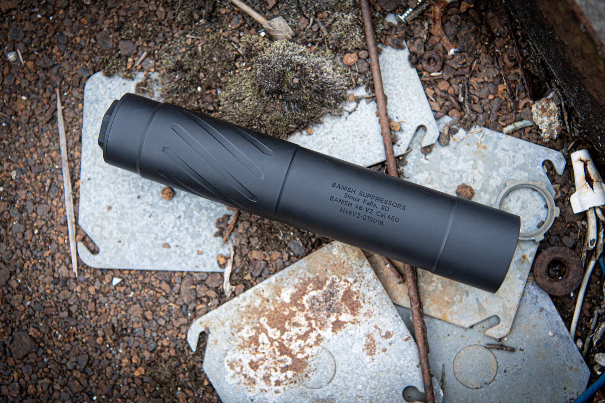 Black rifle suppressor resting on weathered metal plates and rocky ground outdoors, with visible text markings on the cylinder.