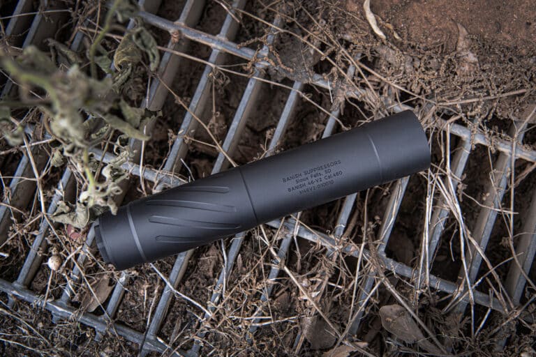 Close-up of a black cylindrical gun suppressor lying on dirt and a metal grate, with dry vegetation around it.