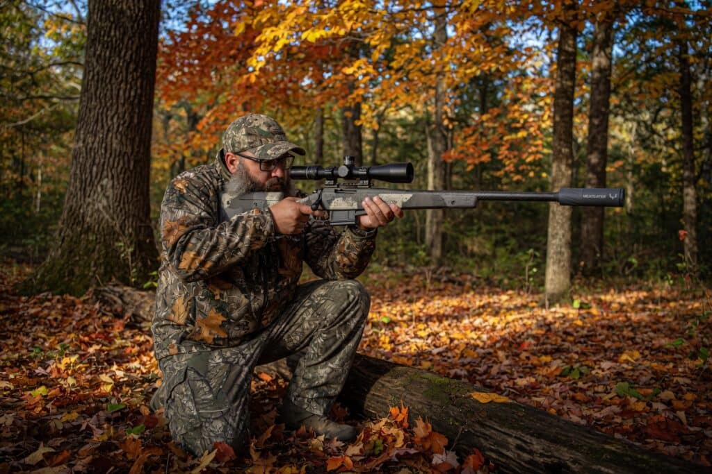 Camouflage-clad hunter crouches on a log in a leaf-strewn autumn forest, aiming a scoped rifle.