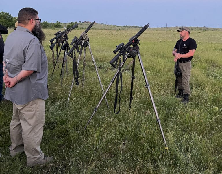 Two men stand in a grassy field beside several tripod-mounted rifles, one with a gray shirt and beard and the other in a black t-shirt, looking on.