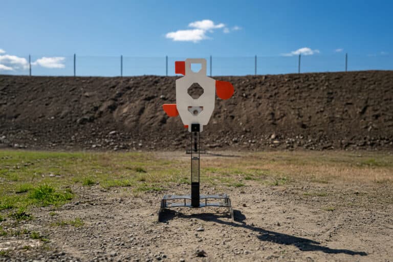 Metal outdoor shooting target on a stand in a dirt field with a blue sky and fence in the background.