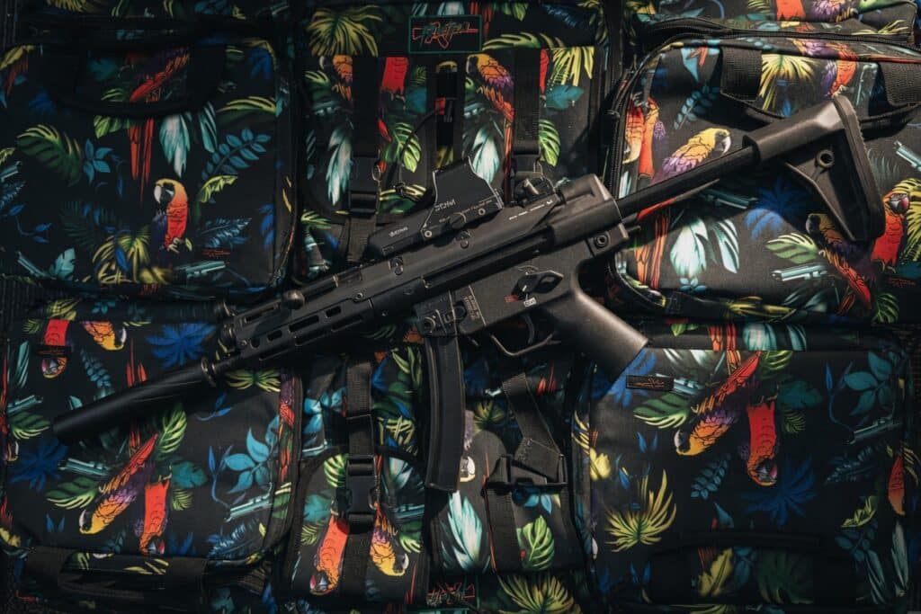 Black assault rifle laid across a tropical-pattern backpack with parrots in bright colors.