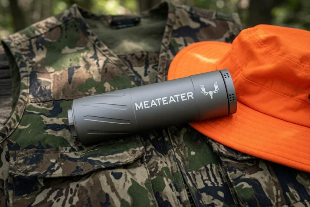 Gray cylindrical Meateater-branded outdoor accessory lying on camouflage jacket with an orange hat nearby outdoors.
