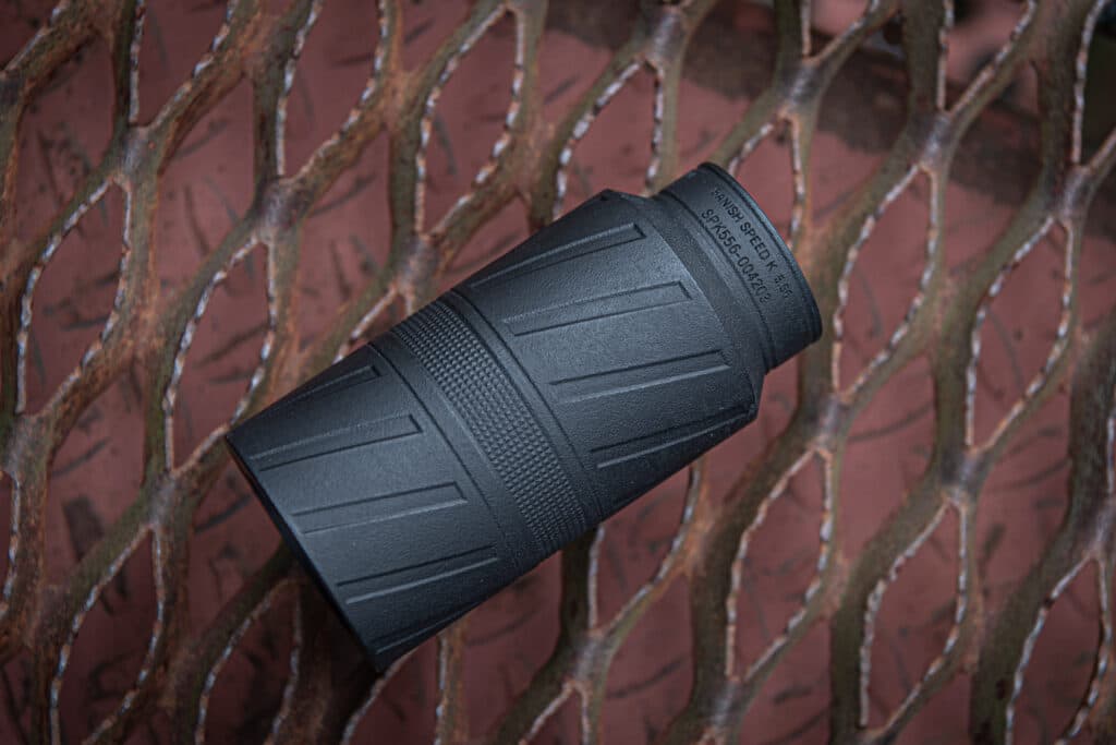 Close-up of a black binocular/monocular device with a textured grip against a rusted chain-link fence background.