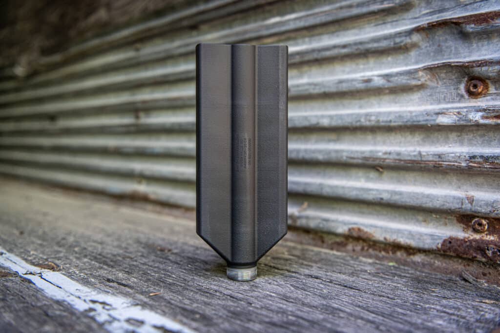 Close-up of a matte black plastic component with two vertical ridges mounted on a threaded metal base, against a weathered corrugated metal background.”,