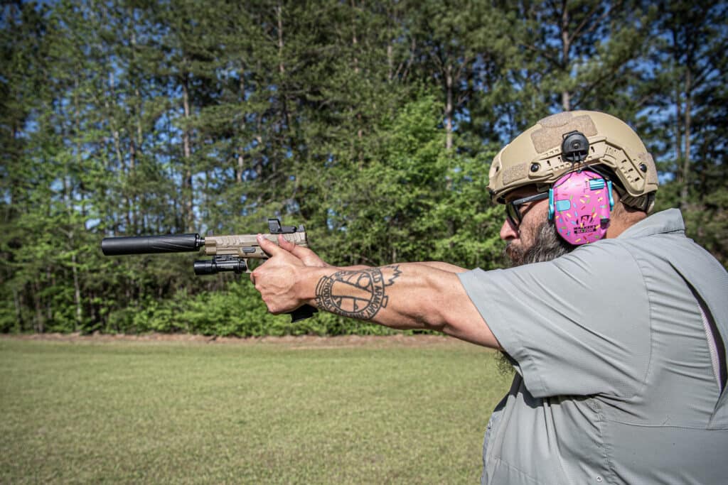 Man in tactical helmet and pink patterned ear protection aims a rifle outdoors, with a tattooed forearm visible.