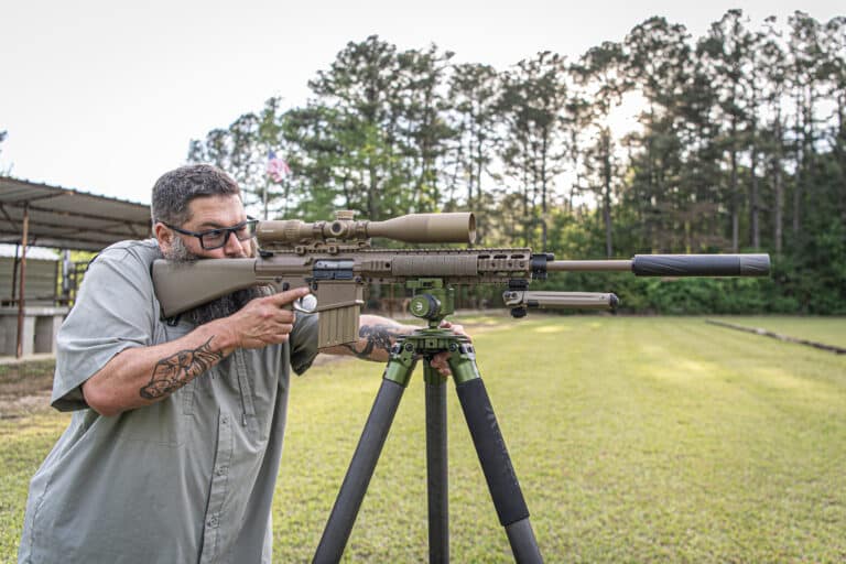 Man aiming a tan rifle with a large scope mounted on a bipod outdoors in a grassy area.