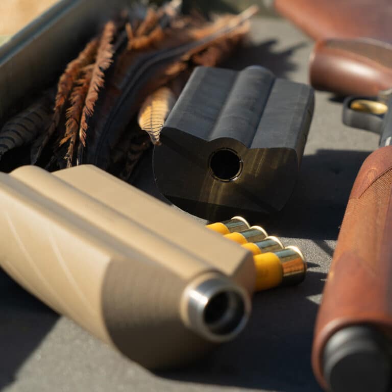 Assorted firearm components on a work surface: a tan rifle stock, a black muzzle/receiver part, and a row of yellow shotgun shells nearby.