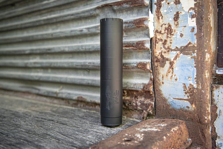 Black cylindrical vape device standing upright on a weathered wooden surface with a rusted, peeling metal backdrop behind it.
