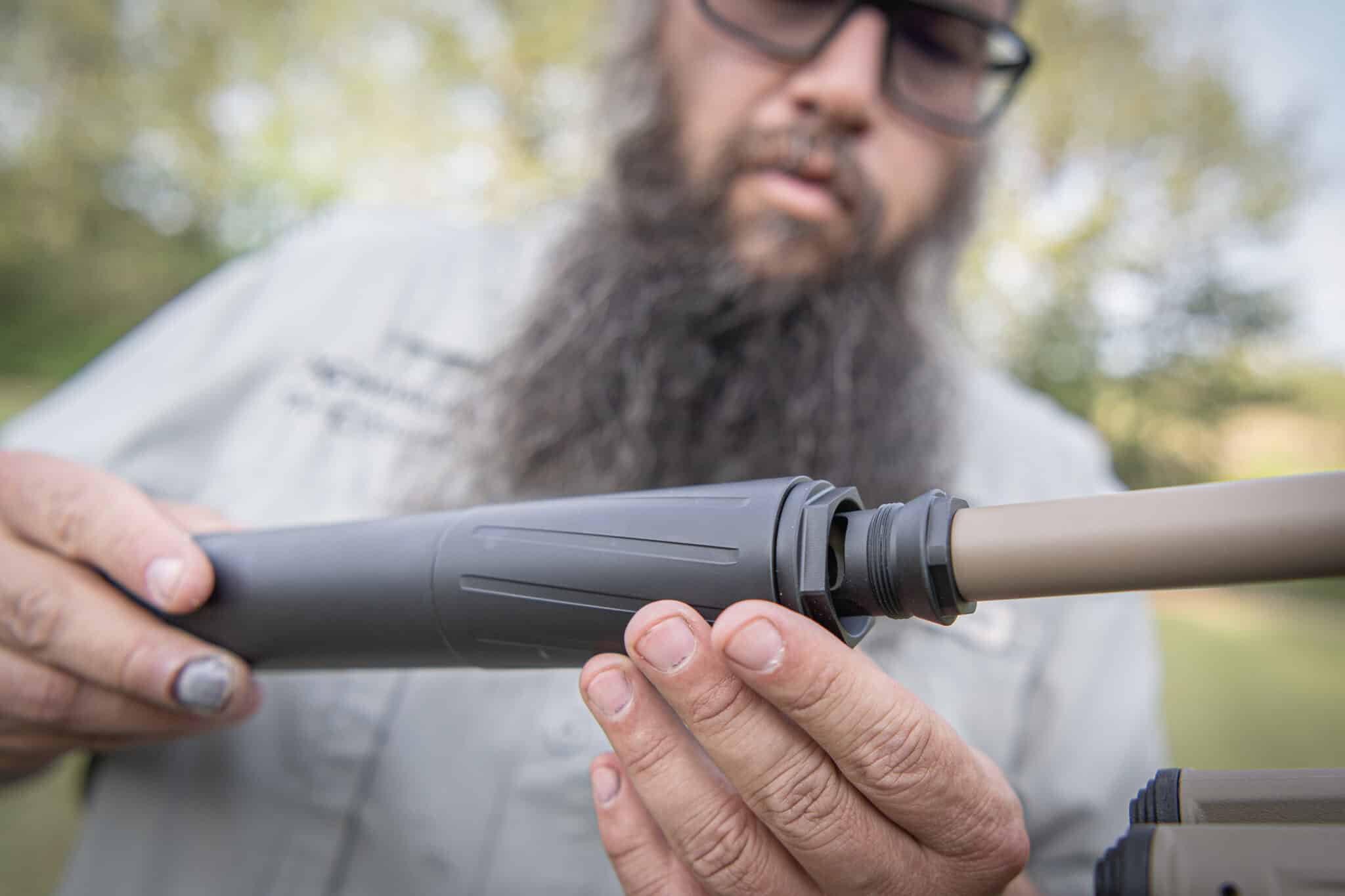 Bearded man wearing glasses holds a rifle-like firearm, assembling it outdoors (close-up).
