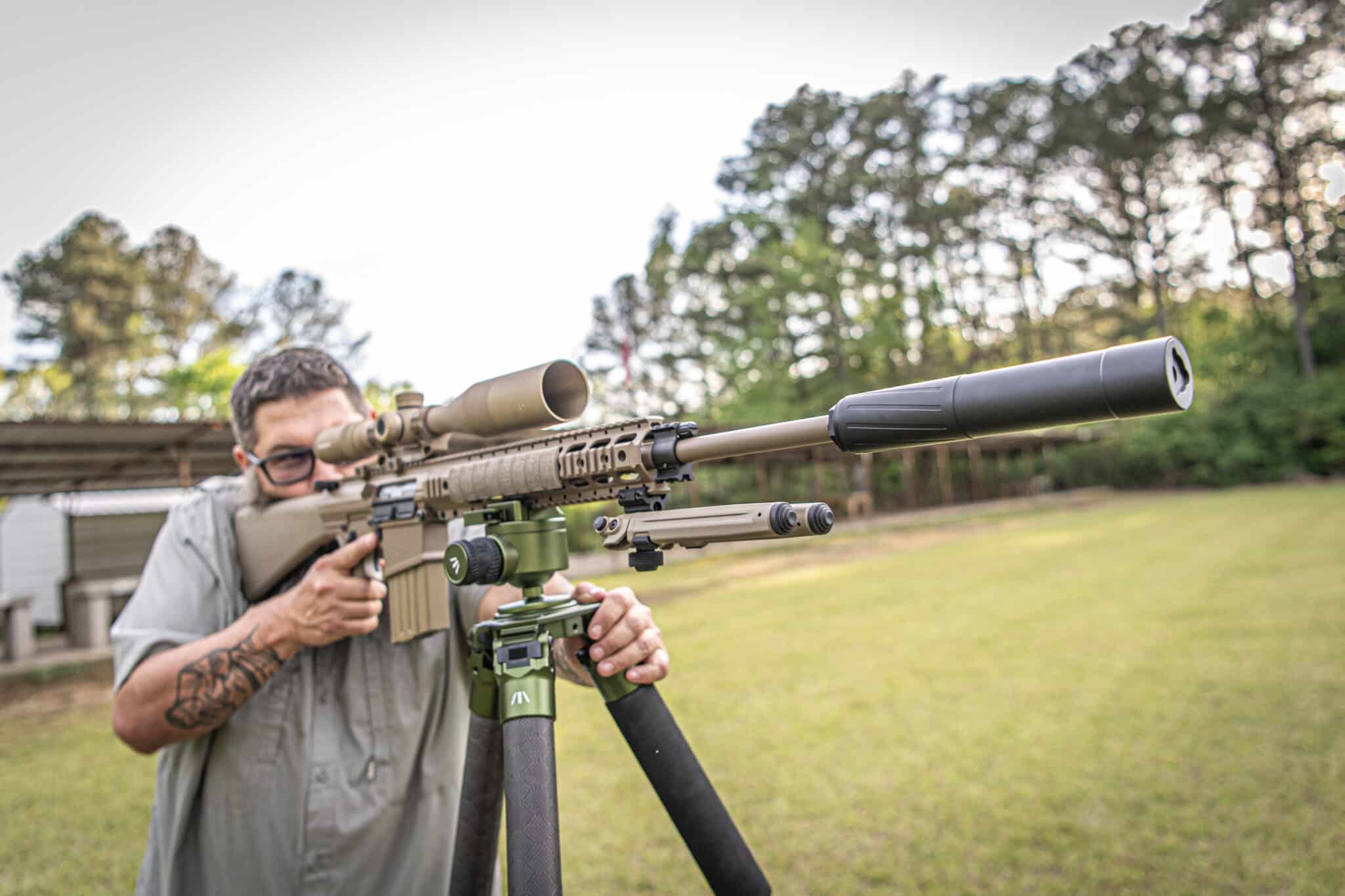 Man aiming a scoped sniper rifle outdoors, close-up on the rifle and shooter’s hands and glasses of the shooter.
