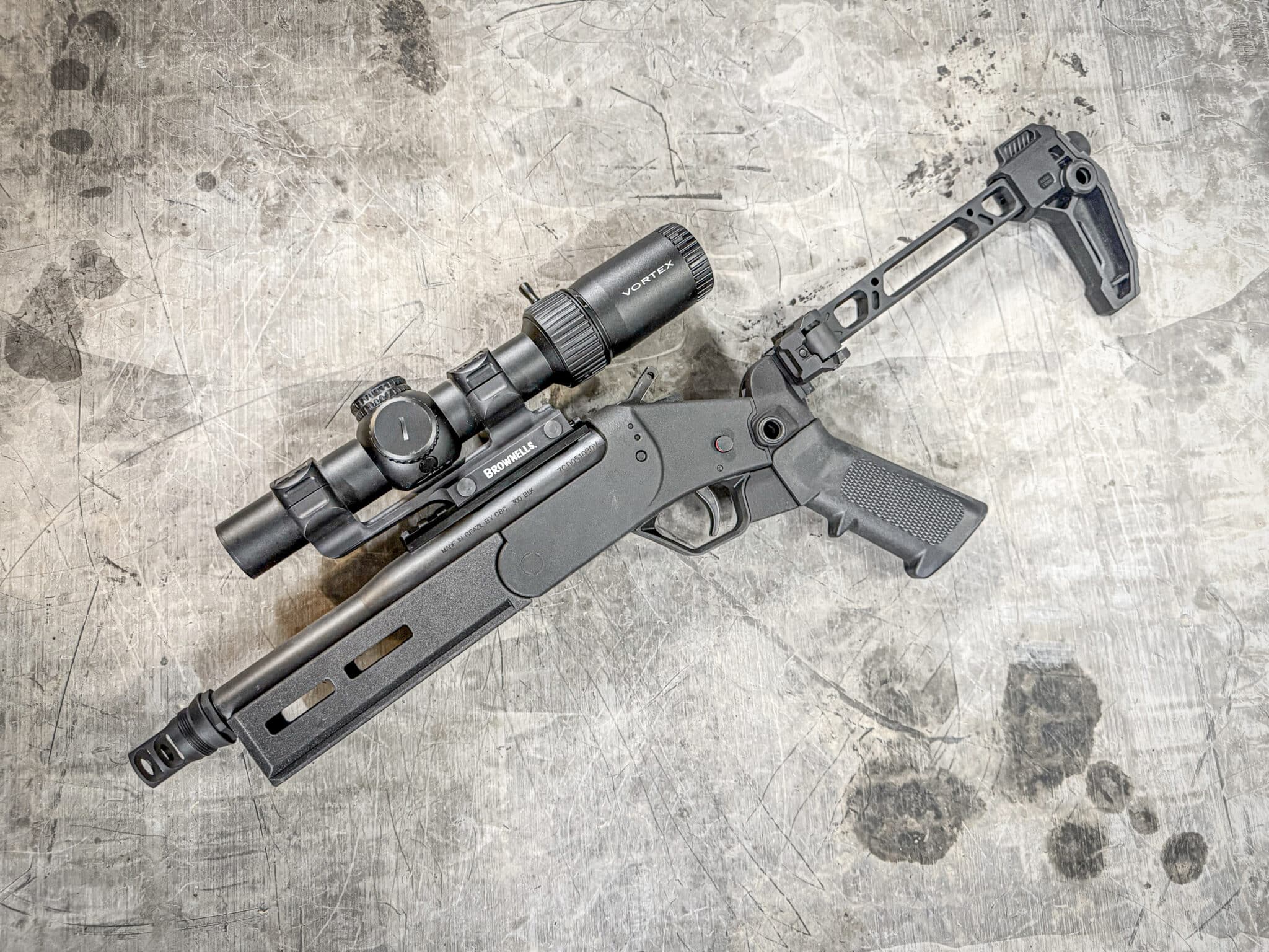 Close-up of a rifle with a mounted optic sitting on a scratched concrete floor outside a workshop setting.