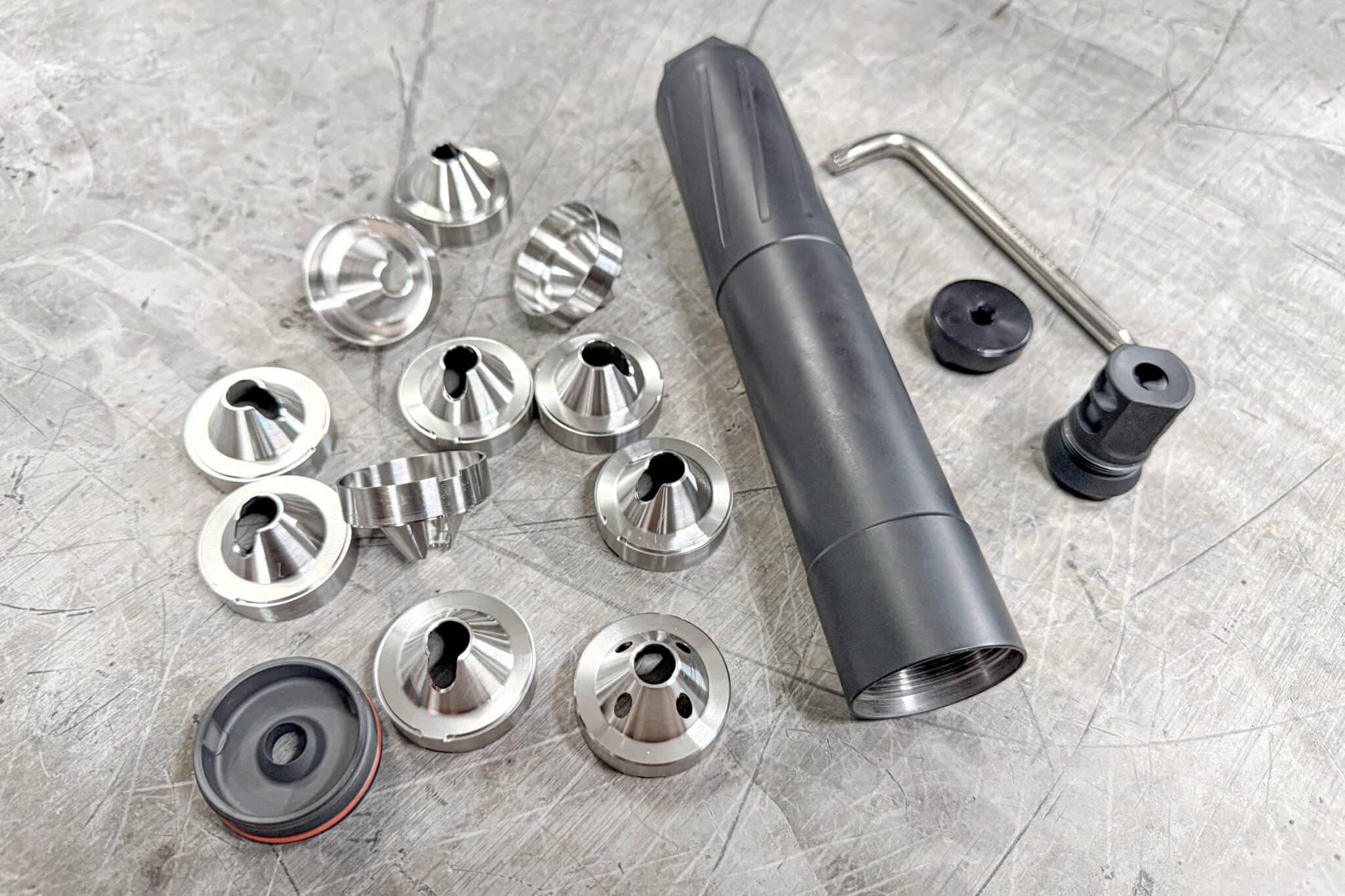 Assorted small metal conical ferrules and spacers scattered on a gray workshop surface with a black cylindrical tool nearby.