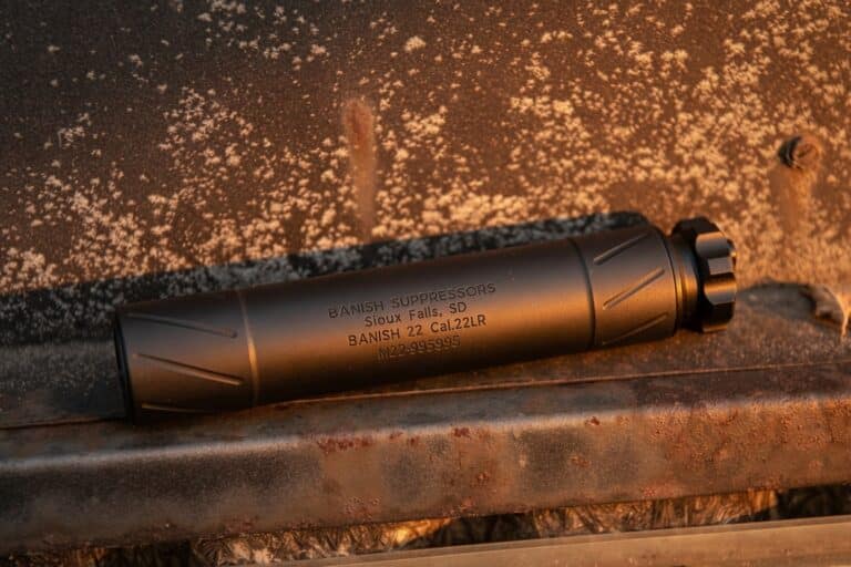 Cylindrical gun suppressor with engraved text resting on a dusty, rusted metal surface under warm light.