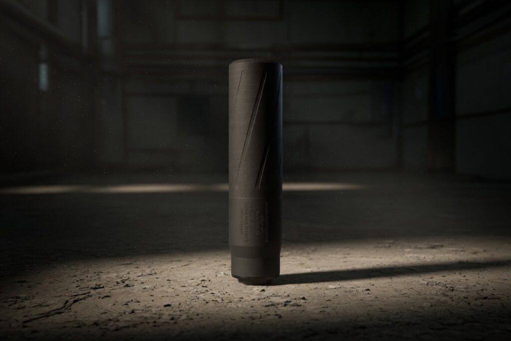 A tall black cylindrical device stands upright on a dusty warehouse floor, lit by a single spotlight in a dark, industrial space.