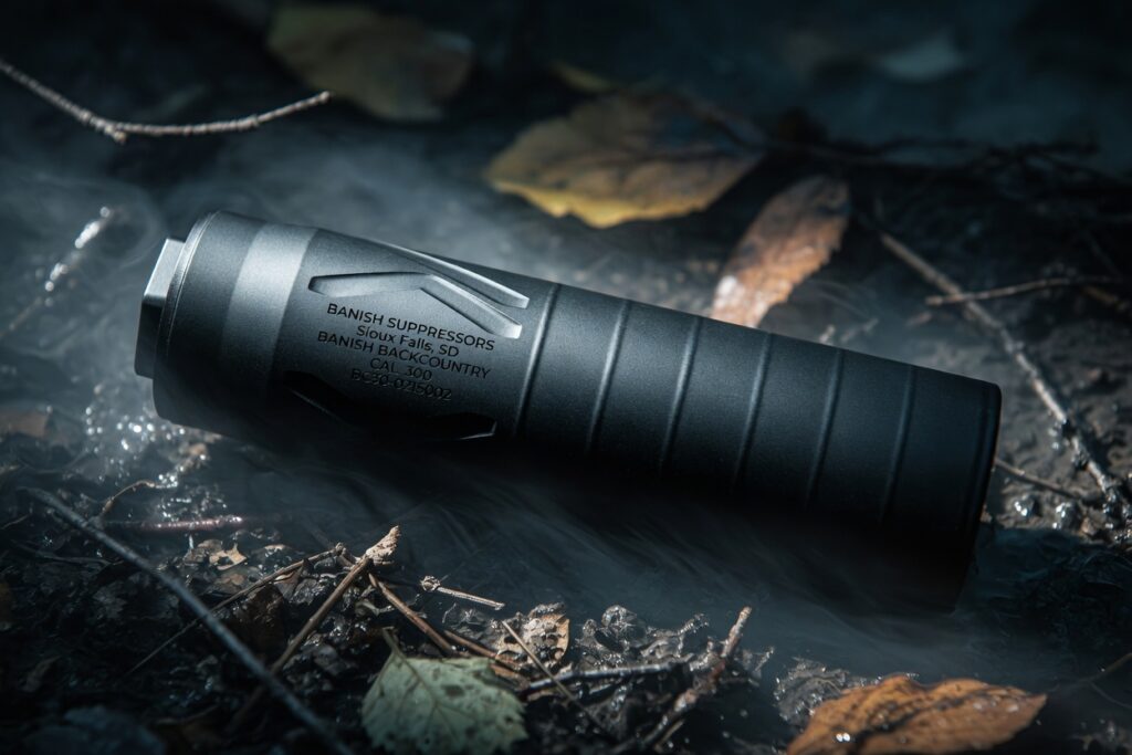 Black firearm suppressor lying on a damp forest floor with leaves and fog.