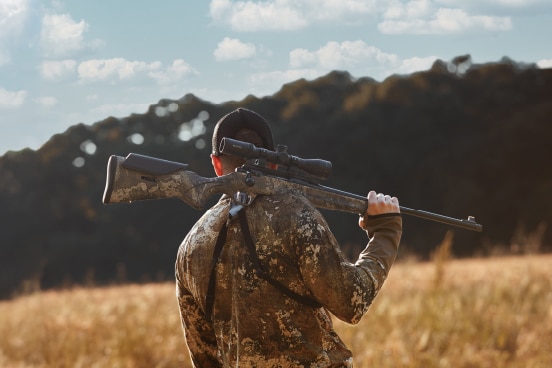 Person in camouflage carrying a scoped rifle over their shoulders in a grassy field with trees in the background, seen from behind.