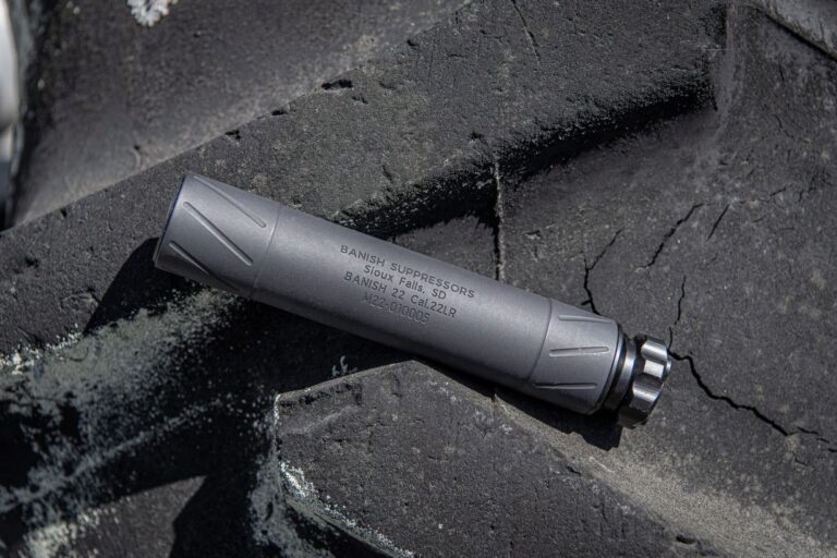 Close-up of a gray tactical pen resting on a dark, rough rock surface; engraving partially visible on the pen's body.