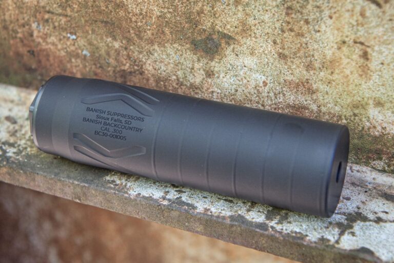 Close-up of a black firearm suppressor lying on a rusted metal surface, with legible markings: BANISH SUPPRESSORS, CAL. .300, BC30-001005