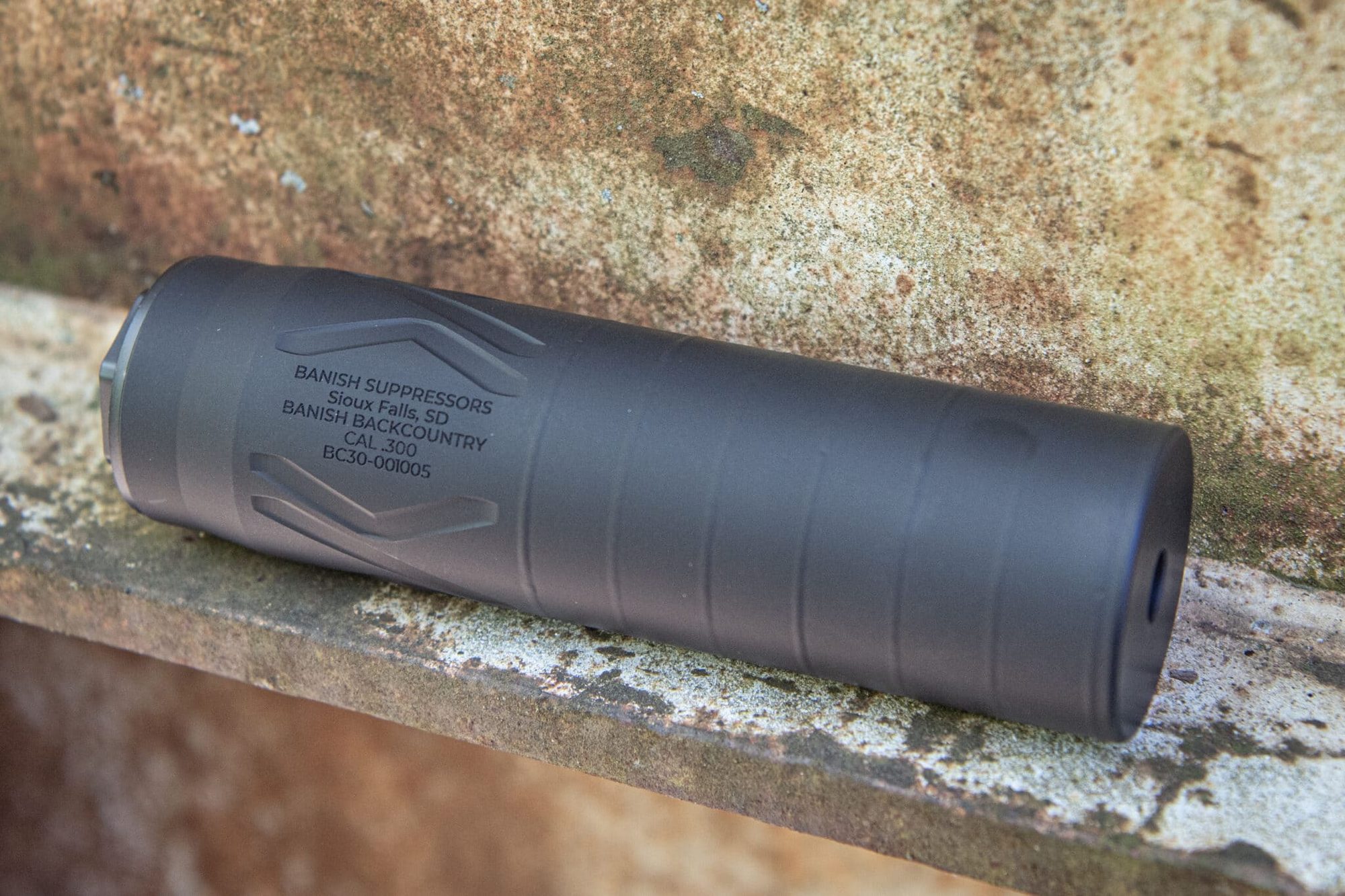 Close-up of a black firearm suppressor lying on a rusted metal surface, with legible markings: BANISH SUPPRESSORS, CAL. .300, BC30-001005
