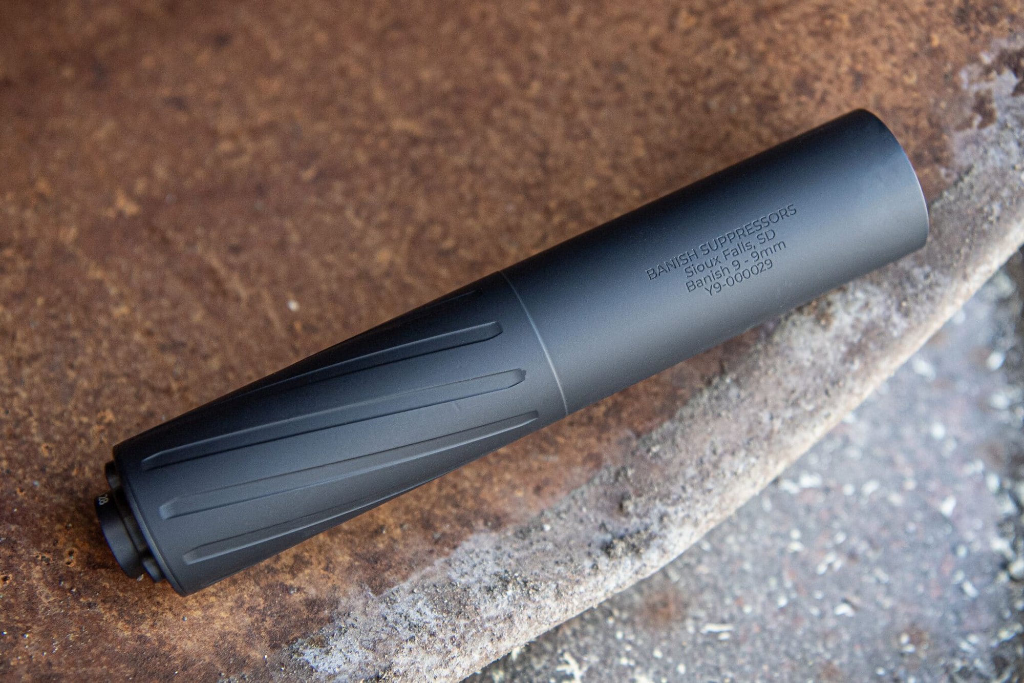 Black suppressor resting on a rusted surface, with engraved text including 'BANISH SUPPRESSORS' and location details Sioux Falls, SD