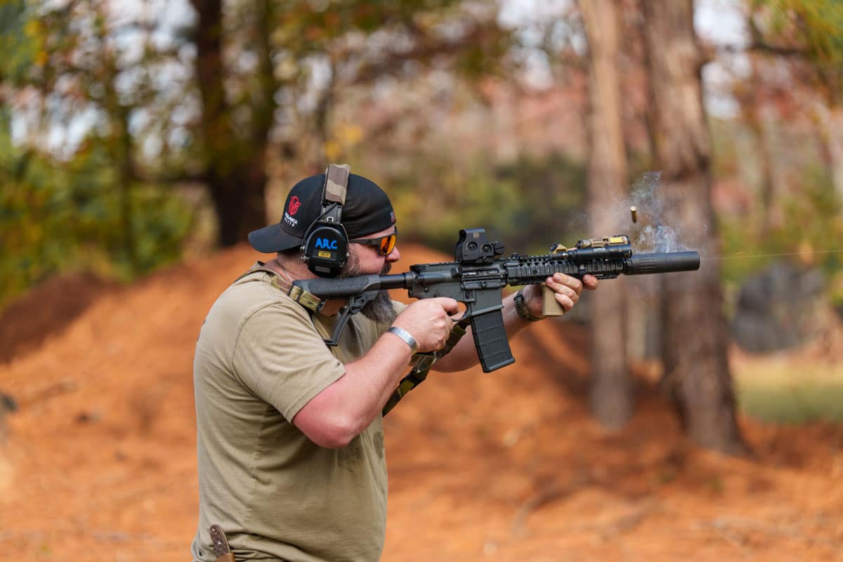 Man in olive shirt firing a black rifle outdoors, wearing hearing protection and sunglasses in a wooded area