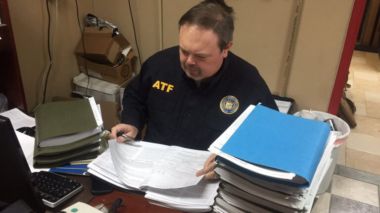 ATF agent in uniform reviews piles of documents at a cluttered desk with binders and folders present
