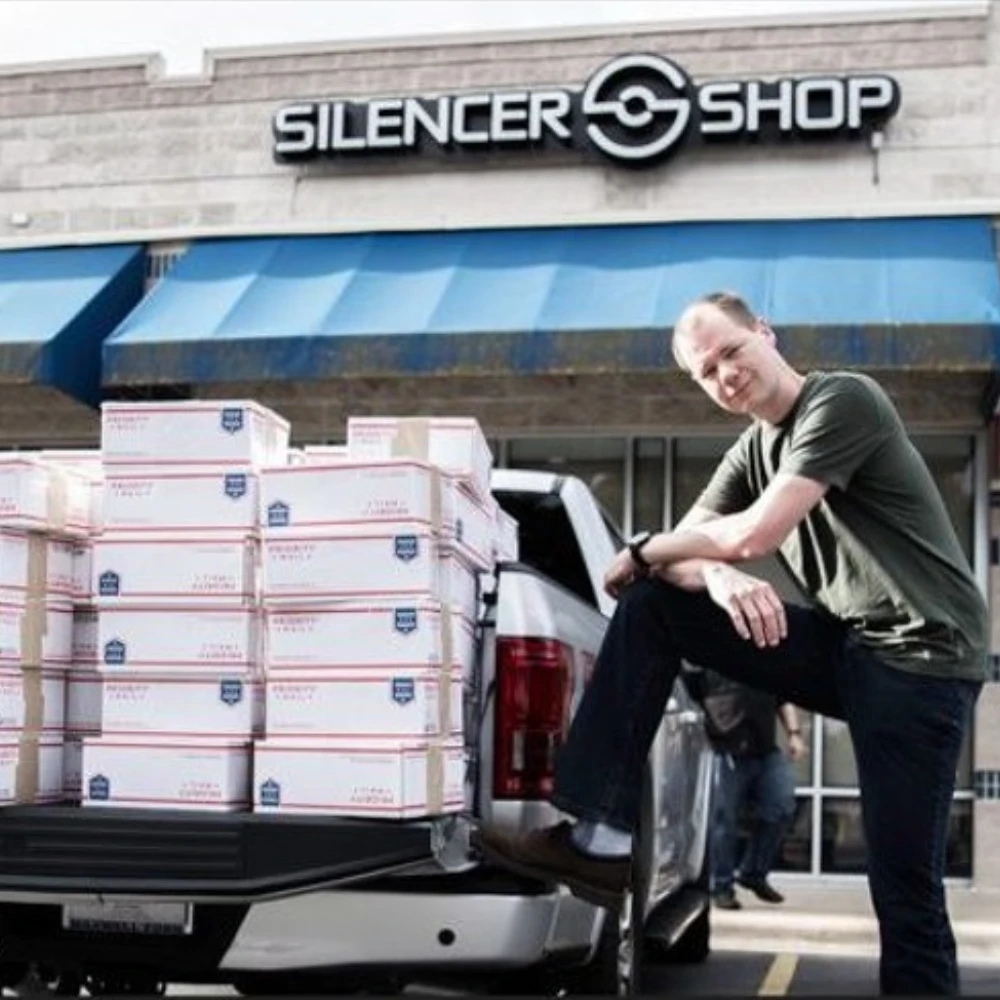 Placeholder_Image_54_png - PopularSuppressors Man leaning on a pickup truck loaded with stacked USPS boxes outside Silencer Shop storefront.