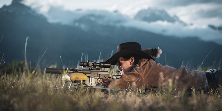 Person wearing a black hat lying prone in a field, aiming a scoped rifle toward the hills in the distance. Striking mountains form the backdrop.