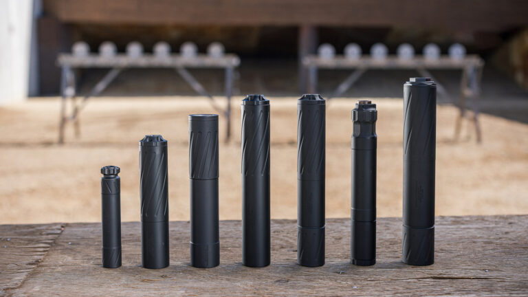 Line of black firearm suppressors standing upright on a wooden table at an outdoor shooting range in the background.